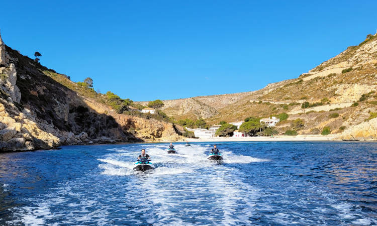 Tres motos de agua navegando en una cala con casas al fondo.